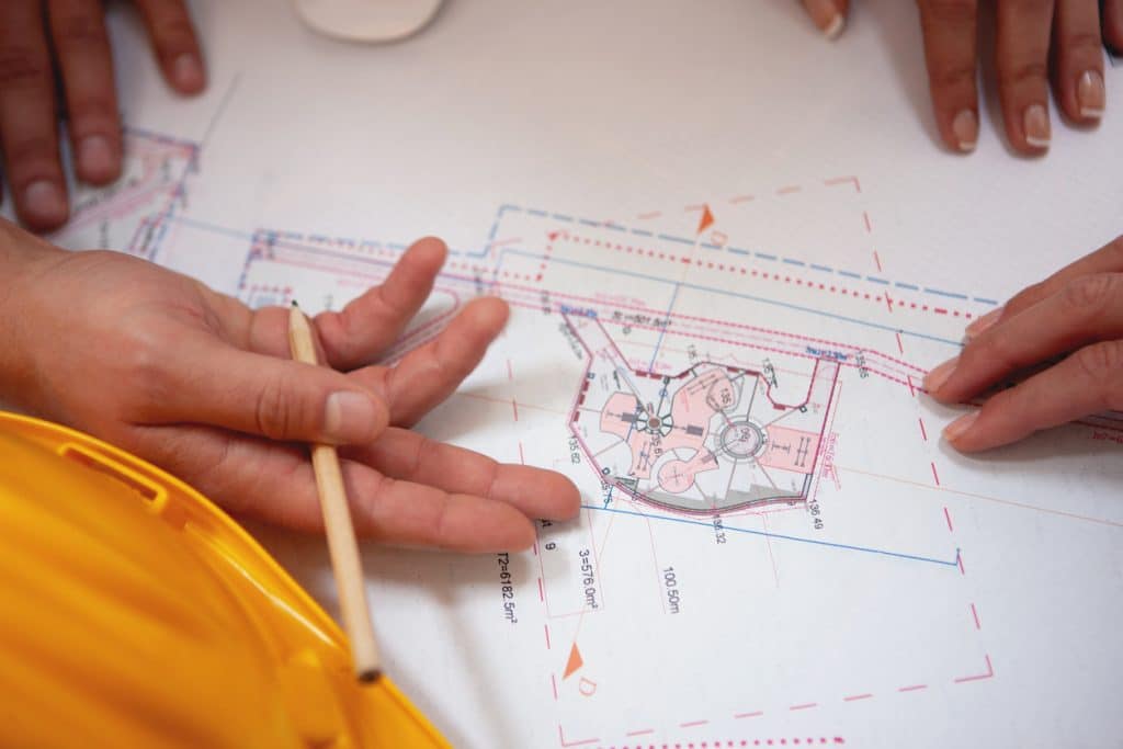 Two hands, one holding a pencil and a blueprint, are positioned to imply discussion. A hardhat is visible.