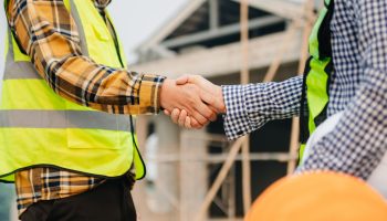Two Men In Safety Vests Shaking Hands At A Commercial Construction Site.