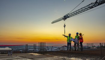 Three Contractors Stand Atop A Building At Dawn, Framed By A Large Crane, Pointing And “looking Toward Tomorrow.”