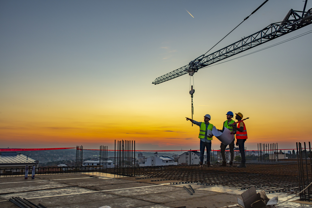 Three contractors stand atop a building at dawn, framed by a large crane, pointing and “looking toward tomorrow.”