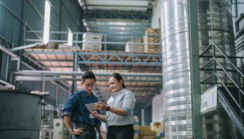 An Image Of Two People Looking At A Tablet In A Factory Setting, Surrounded By Storage Such As Pallet Racks.