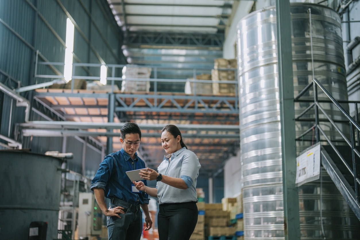 An image of two people looking at a tablet in a factory setting, surrounded by storage such as pallet racks.