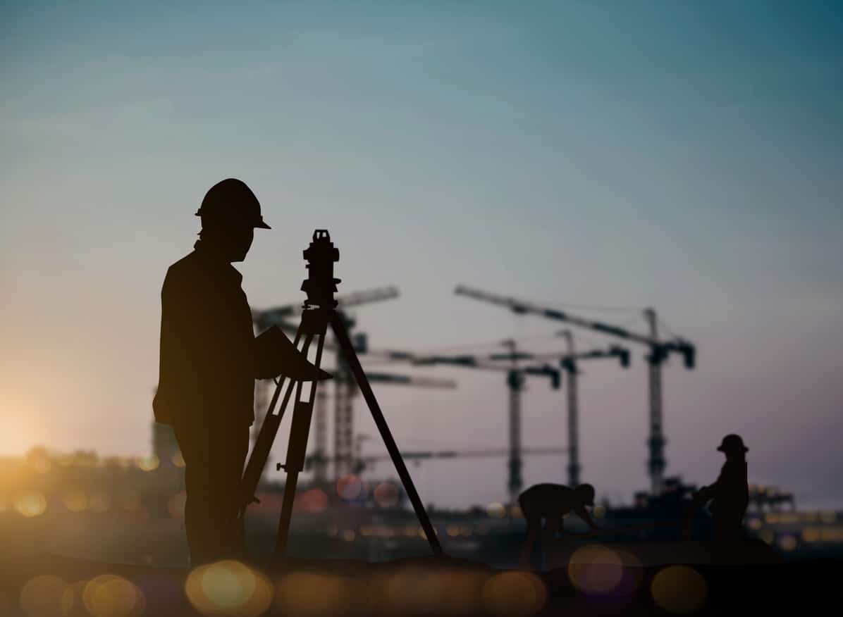 An image of a surveyor taking measurements in the foreground of a large construction site.