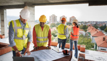 A Team Of People In Construction Vests Discuss A Blueprint At A Construction Site.