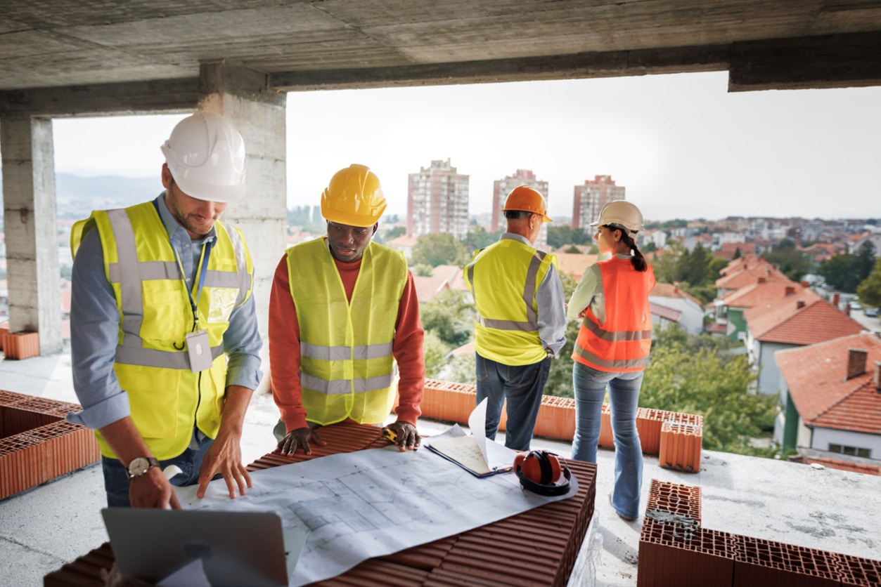 A team of people in construction vests discuss a blueprint at a construction site.